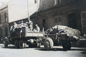 Camion avec des soldats armés, tractant une remorque pleine de bâches de camouflages, stationnant devant des maisons.