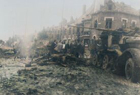 Véhicule militaire à chenille arrière munie d'un canon totalement calciné à côté de celui-ci des civils, le sol est jonché de débris métalliques et de pierres au croisement de l'avenue de Valenciennes et l'avenue du Quesnoy