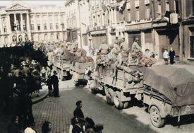 Défilé de camions avec remorques remplis de soldats américains assis qui se trouvent en haut de l'avenue de la victoire entourés de badauds partant en direction de l'hôtel de ville