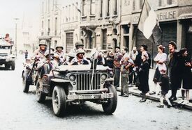 Des soldats américains dans une Jeep avec remorque remontant l'avenue de la Victoire et sur le côté du magasin Ornement d'église des badauds et un drapeau français